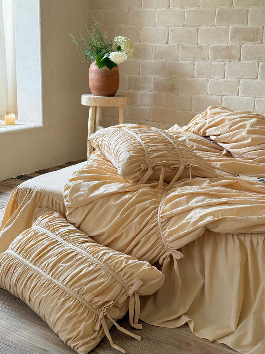 Ruched cotton pillowcase with piped channels and bow ties beside a cream ruched duvet cover and ruffled bed skirt