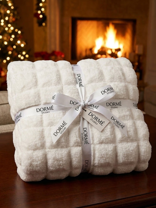 White Marshmallow Ultra Plush Blanket with a grid texture, folded and tied with a DORMÉ Studio satin ribbon, set on a wooden table in front of a lit fireplace and Christmas tree.