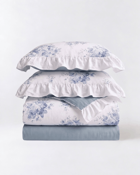 A stack of bedding set including two white pillowcases with blue floral toile de Jouy patterns and ruffled edges, a matching folded sheet with the same floral print, and a solid dusty blue folded sheet underneath, all on a clean white background.