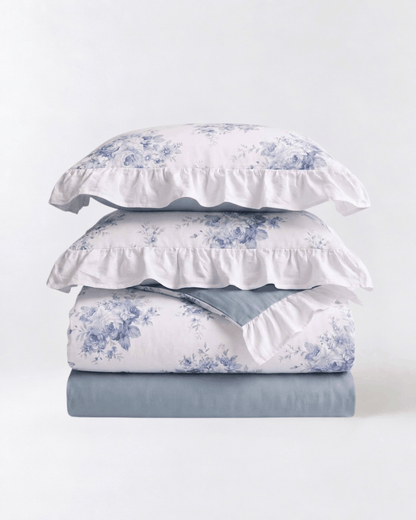 A stack of bedding set including two white pillowcases with blue floral toile de Jouy patterns and ruffled edges, a matching folded sheet with the same floral print, and a solid dusty blue folded sheet underneath, all on a clean white background.