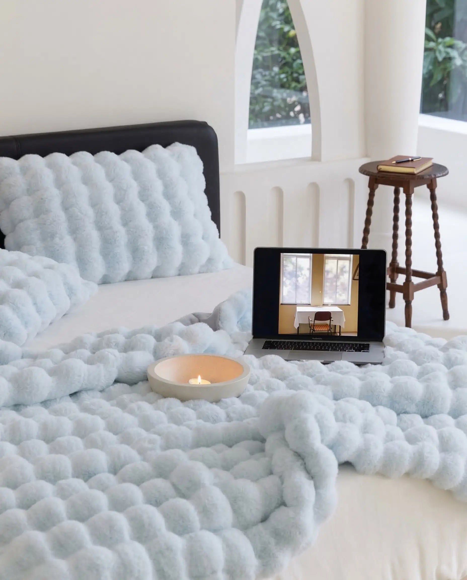 Bed styled with DORMÉ Studio Marshmallow Ultra Plush Blanket in blue plush fabric with candle and laptop creating cozy bedroom aesthetic.