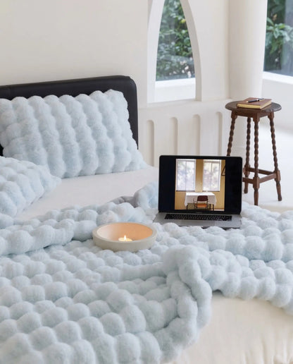 Bed styled with DORMÉ Studio Marshmallow Ultra Plush Blanket in blue plush fabric with candle and laptop creating cozy bedroom aesthetic.