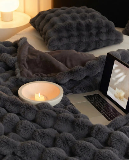 DORMÉ Studio Marshmallow Ultra Plush Blanket in charcoal plush fabric styled on bed with candle and laptop creating cozy bedroom atmosphere.