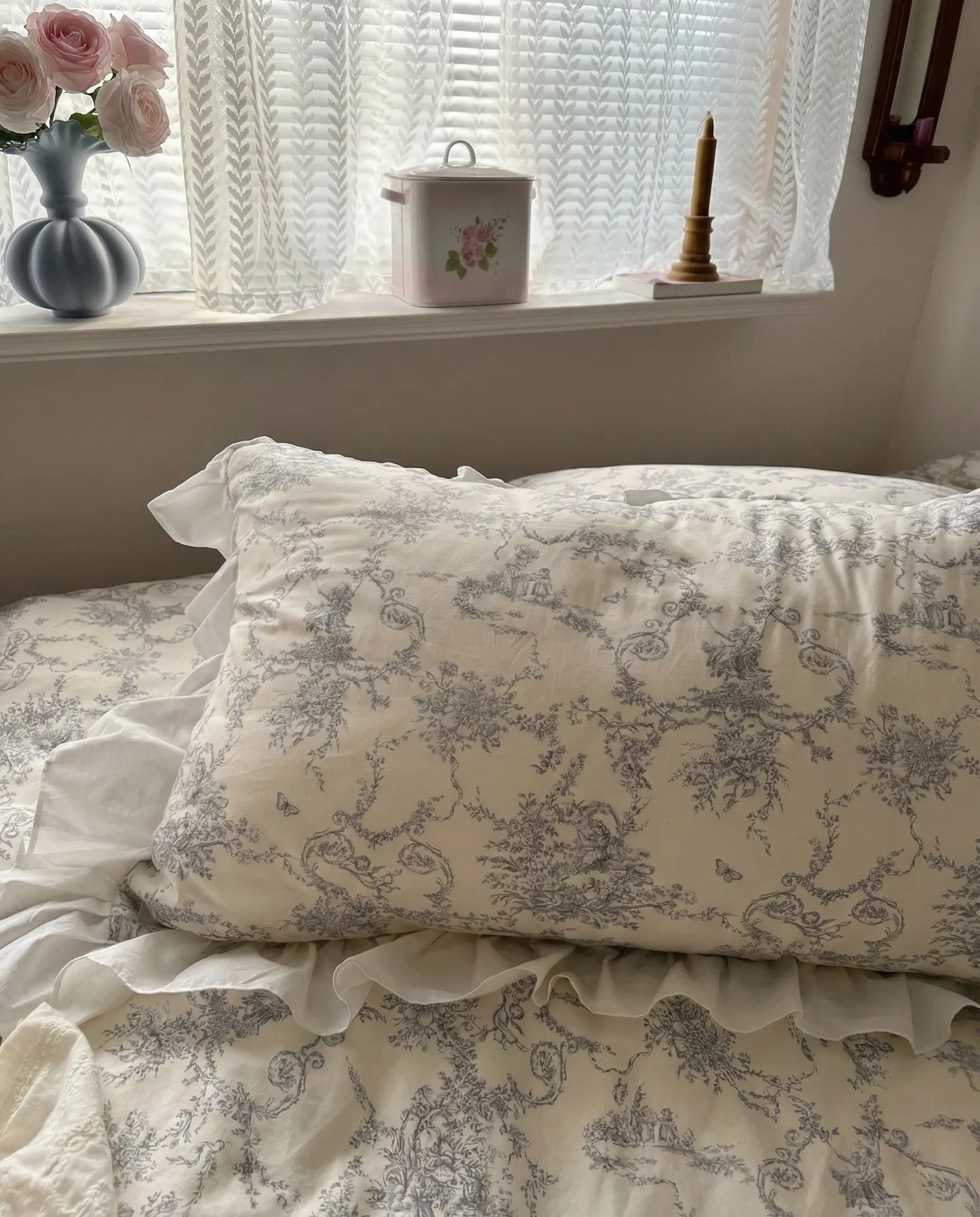 Grey floral pillow sham with ruffled edge resting near a bright window, featuring classic toile print on white bedding, aesthetic bedroom, vintage bedding, floral pillows, white bedding, grey floral, romantic bedroom, cottage bedroom, bedroom decor