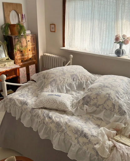 Sunlit bedroom featuring a grey floral ruffled duvet cover on a white metal bed, styled with vintage furniture and soft decor, aesthetic bedroom, vintage bedding, floral duvet, white bedding, grey floral, romantic bedroom, cottage bedroom, bedroom decor