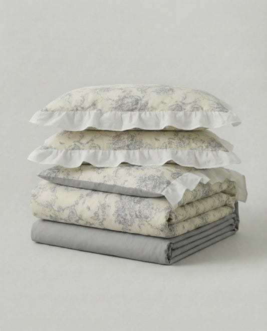 Folded floral duvet cover set with matching ruffled pillowcases in soft ivory and gray, showcasing delicate vintage toile pattern and layered bedding textures