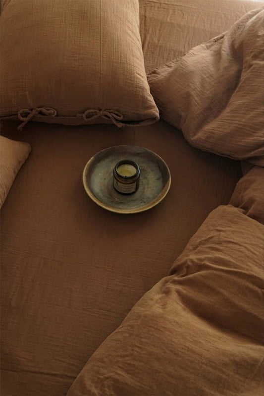 Overhead view of Cinnamon muslin bedding with tie-closure pillowcases and an amber candle on a ceramic plate