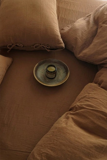 Overhead view of Cinnamon muslin bedding with tie-closure pillowcases and an amber candle on a ceramic plate