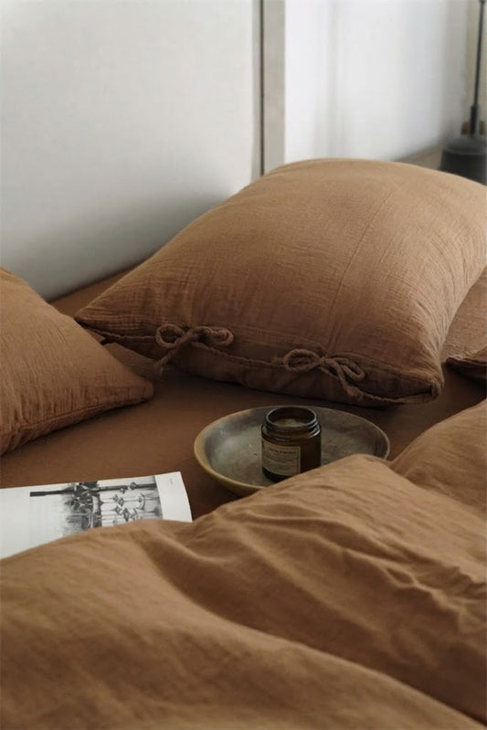 Cinnamon muslin pillowcase with side tie closures beside an amber candle and a magazine on a made bed