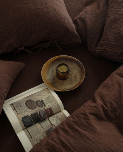 Overhead view of chocolate espresso muslin bedding with tie-closure pillowcases, a candle, and an open book