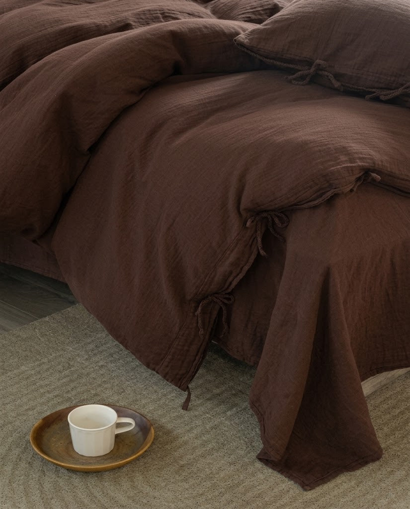 Corner of a chocolate espresso muslin duvet cover with tie closures draping over the bed onto a textured rug