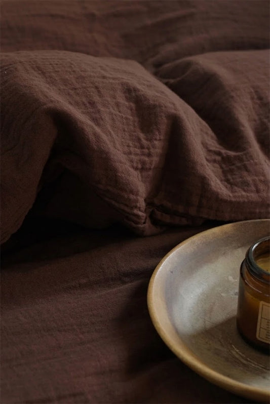 Close-up of crinkled chocolate espresso muslin pillowcase texture beside an amber candle on a ceramic plate