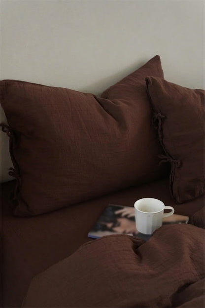Chocolate Espresso muslin pillowcases with side tie closures, a white mug, and a magazine on a made bed