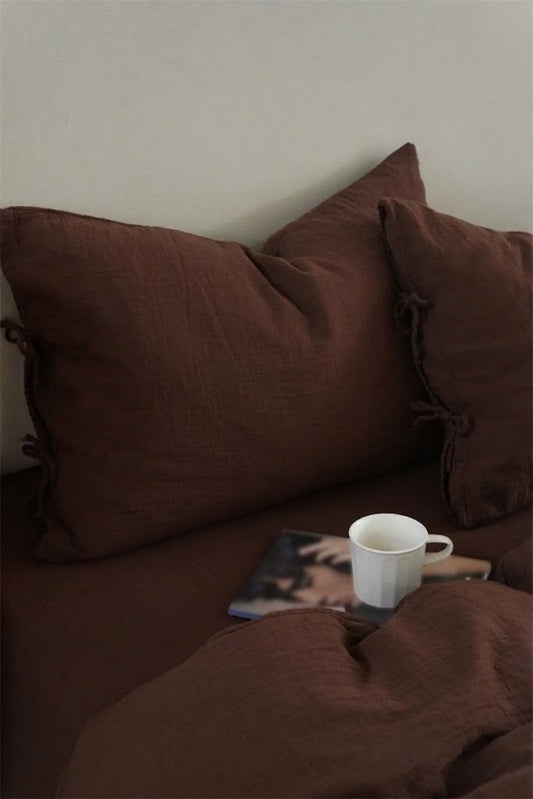Chocolate Espresso muslin pillowcases with side tie closures, a white mug, and a magazine on a made bed
