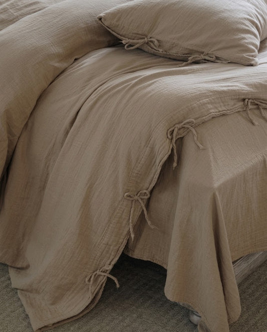 Corner of a Natural Oat muslin cotton duvet cover with bow tie closures draping over the bed onto a textured rug