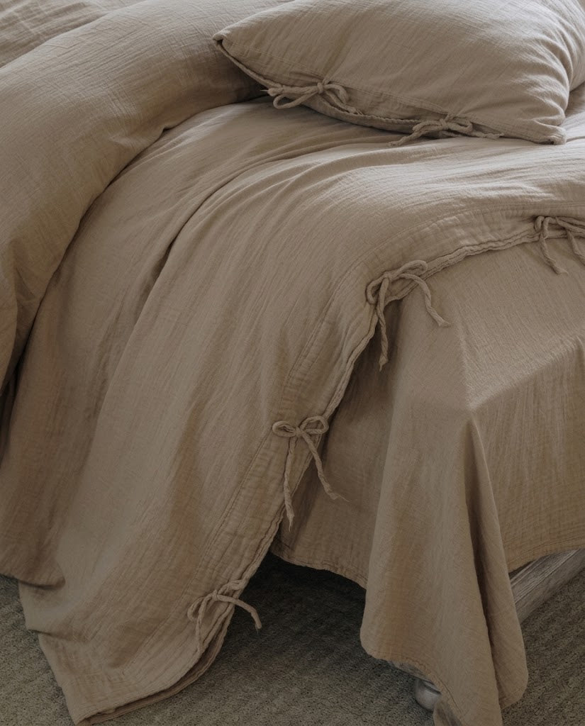 Corner of a Natural Oat muslin cotton duvet cover with bow tie closures draping over the bed onto a textured rug