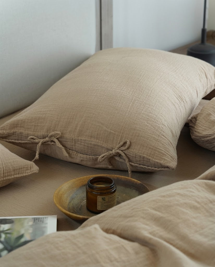 Natural Oat muslin pillowcase with bow tie closures beside an amber candle on a ceramic plate