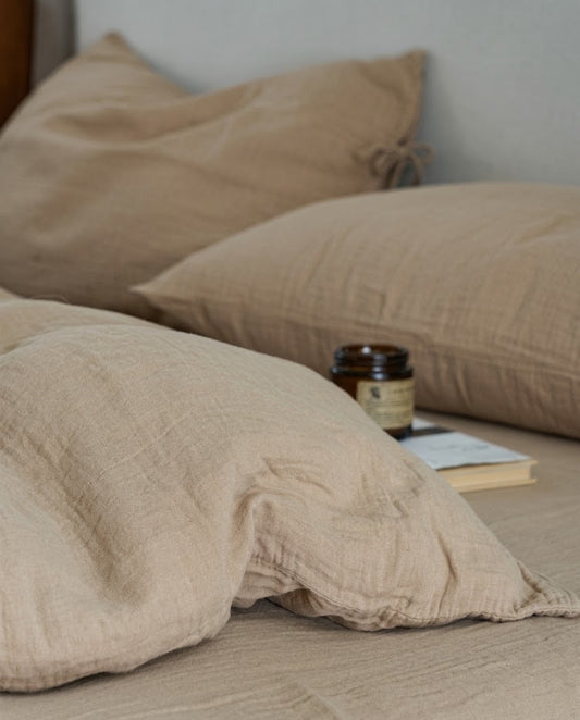 Soft-focus close-up of Natural Oat muslin cotton pillowcases with an amber candle and book on a made bed