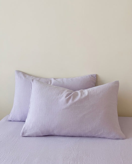 Two lilac muslin cotton double gauze pillowcases on a lilac fitted sheet against a cream wall