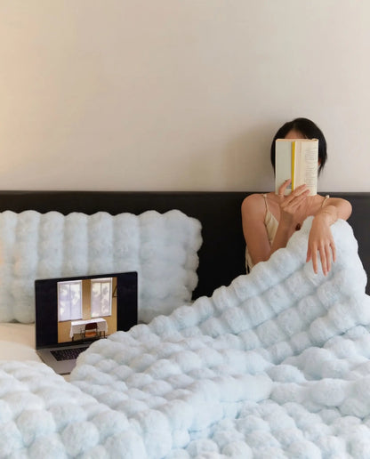 Woman relaxing in bed reading book under DORMÉ Studio Marshmallow Ultra Plush Blanket in soft blue plush fabric.