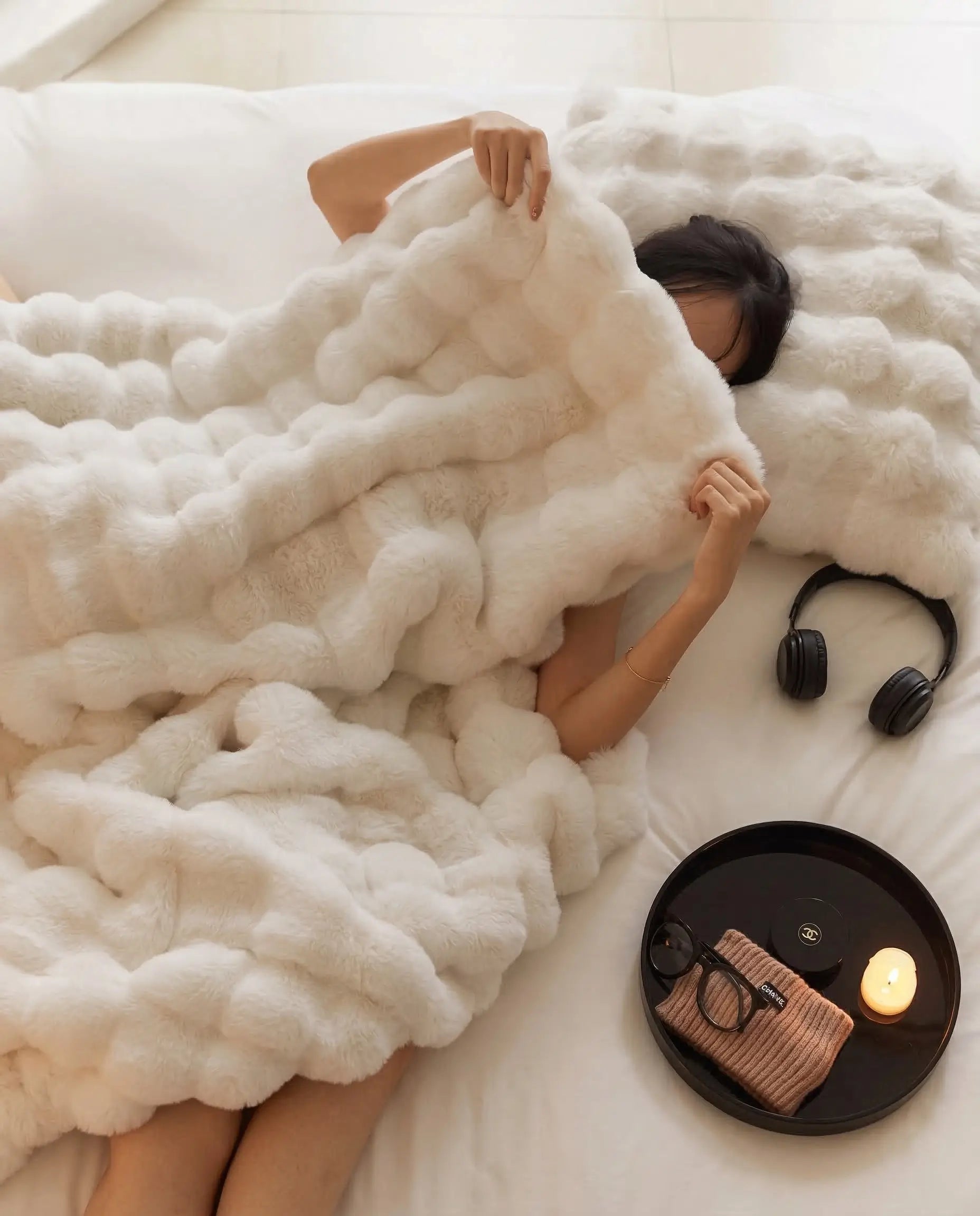 Person wrapped in DORMÉ Studio Marshmallow Ultra Plush Blanket on bed with candle and headphones creating cozy relaxation.