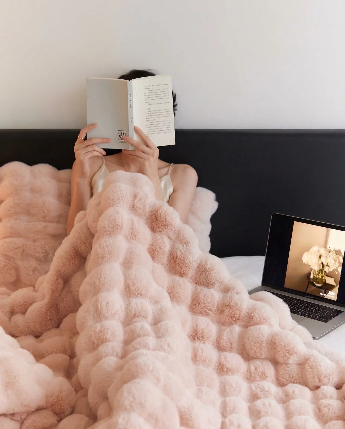 Woman relaxing in bed reading under DORMÉ Studio Marshmallow Ultra Plush Blanket in soft pink plush fabric.