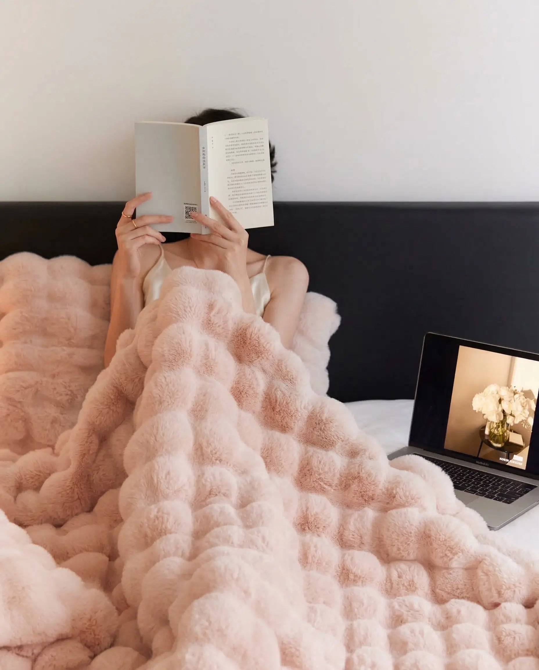Woman relaxing in bed reading under DORMÉ Studio Marshmallow Ultra Plush Blanket in soft pink plush fabric.