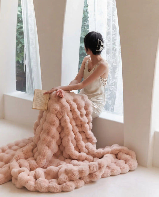 Woman sitting by window wrapped in DORMÉ Studio Marshmallow Ultra Plush Blanket pink plush fabric reading a book. Blossom
