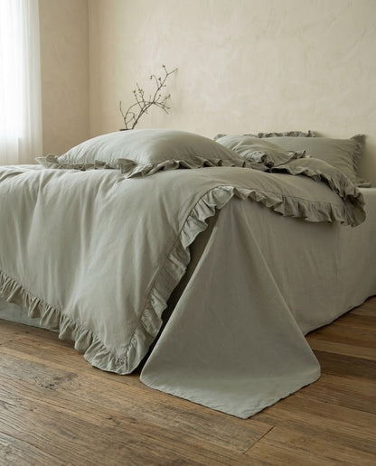 Side view of the Rosalie eucalyptus sage ruffle duvet cover and flat sheet in 100% cotton draping onto a wood floor by a sheer curtain