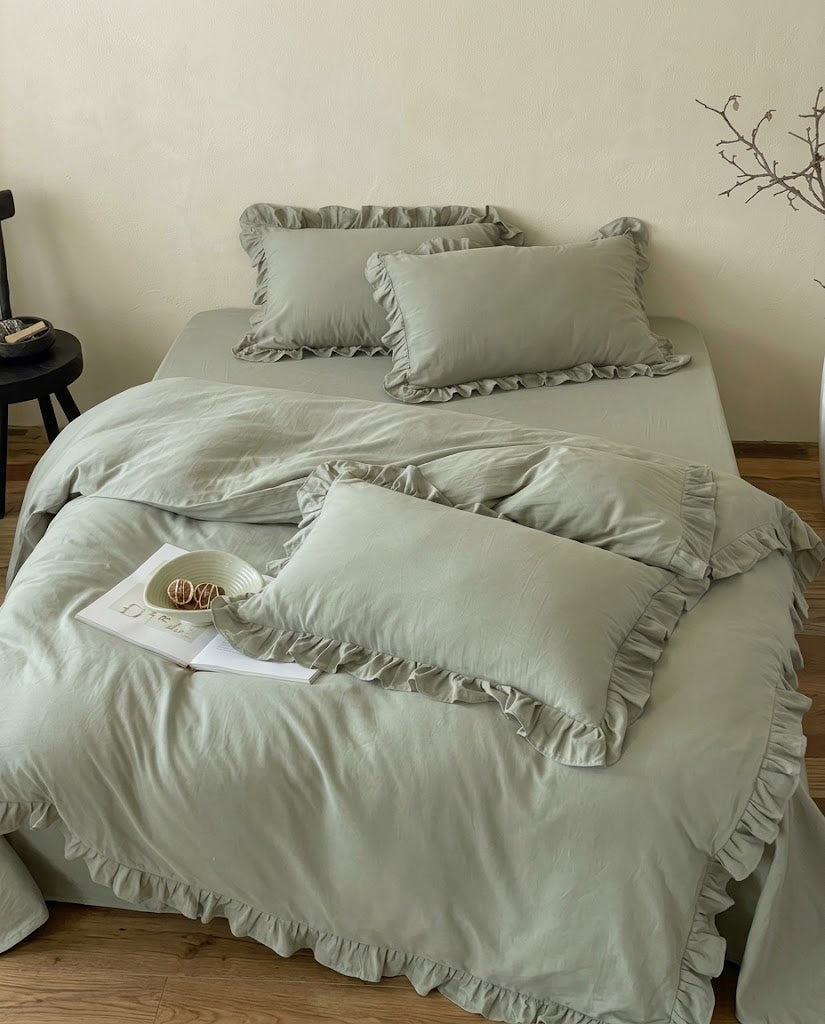 Layered eucalyptus sage ruffle pillow shams on a matching cotton duvet cover from the Rosalie set, with a ceramic bowl and branch
