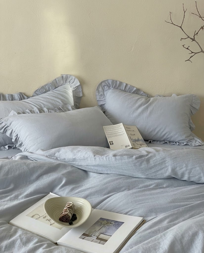 Front view of mist blue ruffle bedding by DORMÉ Studio with layered pillow shams, a rumpled cotton duvet cover and an open art book