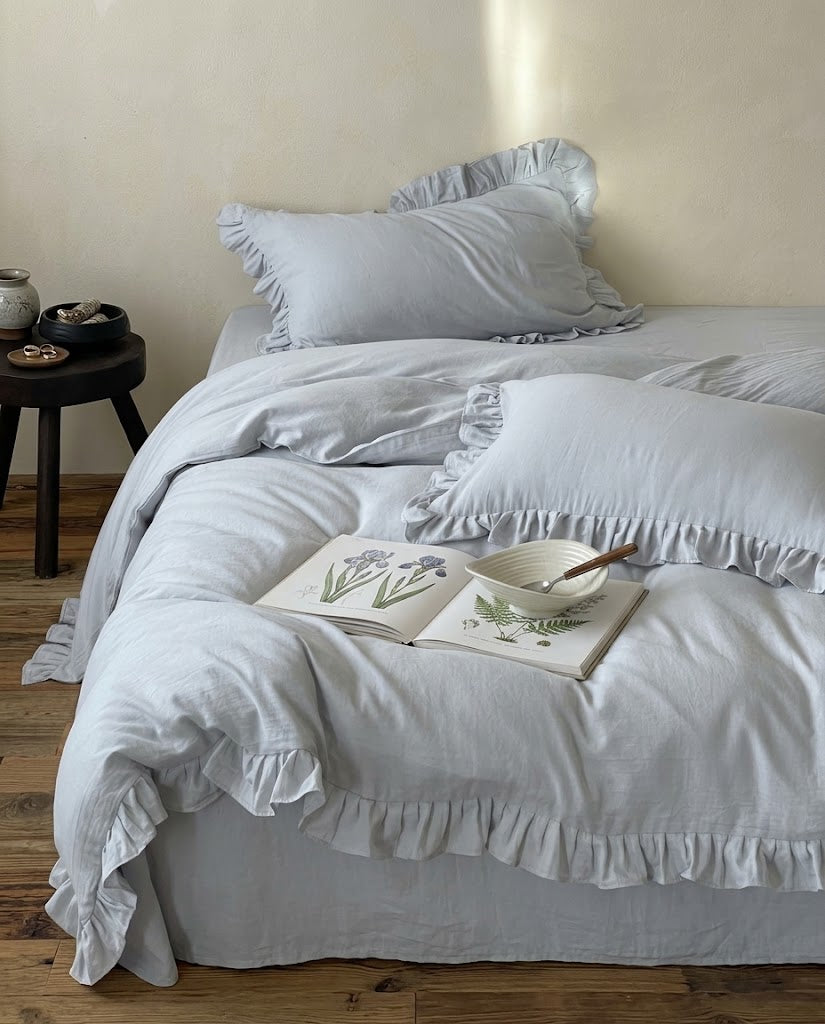 Rosalie mist blue ruffle duvet cover set in 100% cotton on a bed with ruffle pillow shams, an open botanical book and a black side stool