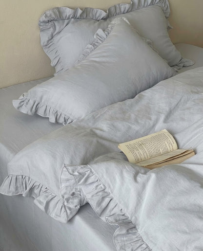 Close-up of the Rosalie mist blue duvet cover and pillow shams showing gathered ruffle trim and soft cotton texture with an open paperback