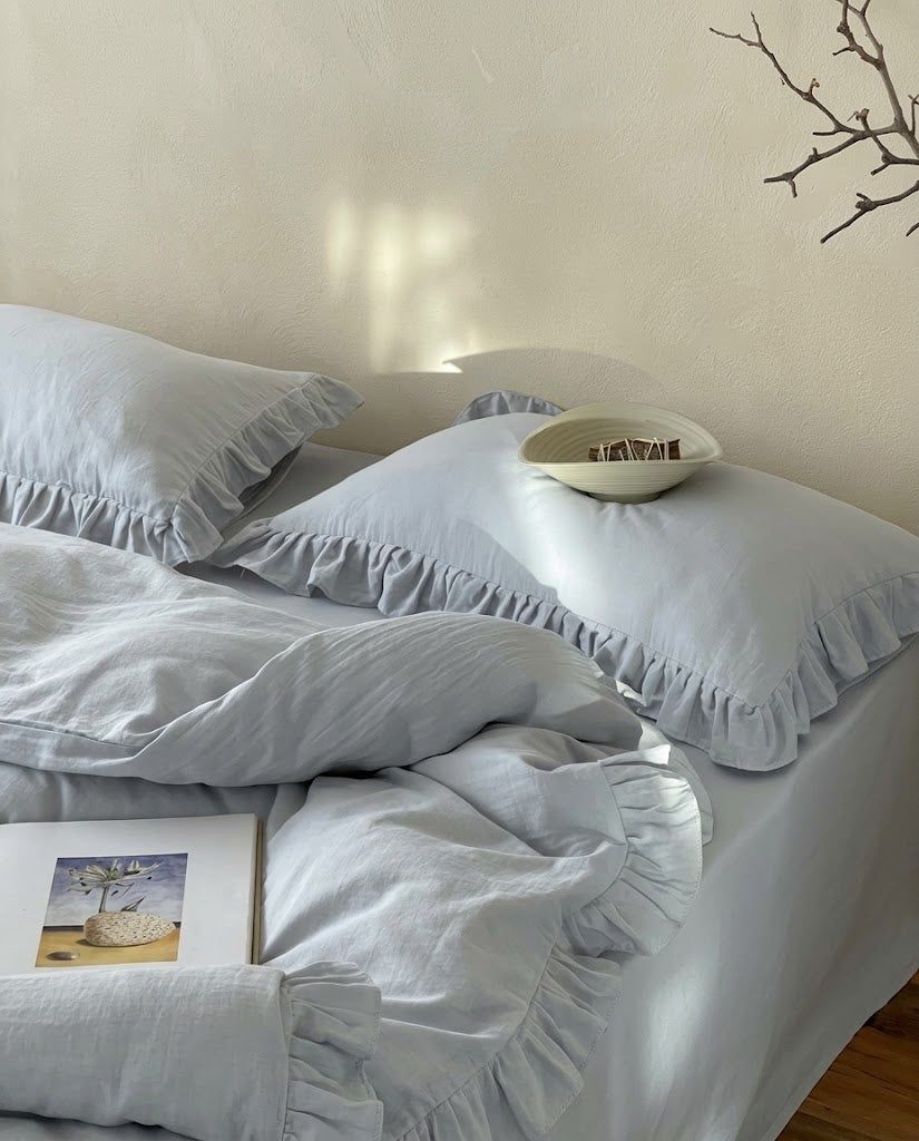 Mist blue ruffle pillow sham and folded duvet cover from the Rosalie set in soft afternoon sunlight with a ceramic bowl and book
