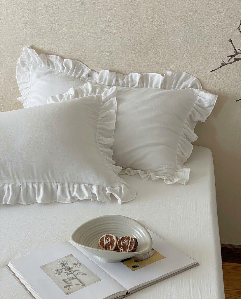 Snow white ruffle pillow shams from the Rosalie duvet cover set by DORMÉ Studio, stacked on a white bed with a ceramic bowl and book