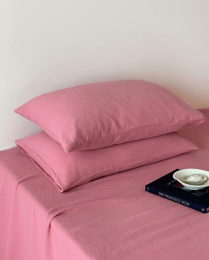 Two Rose muslin cotton double gauze pillowcases stacked on a matching flat sheet with a book and ceramic dish