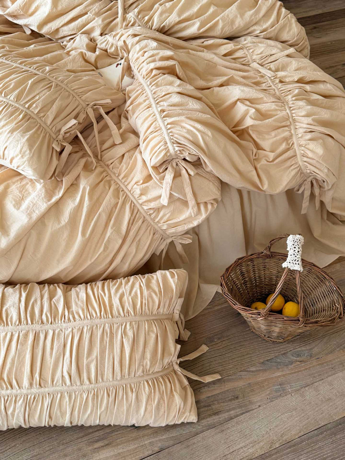 Top-down view of cream washed cotton ruched pillowcase and bow-tied duvet cover with ruffled bed skirt on a wooden floor