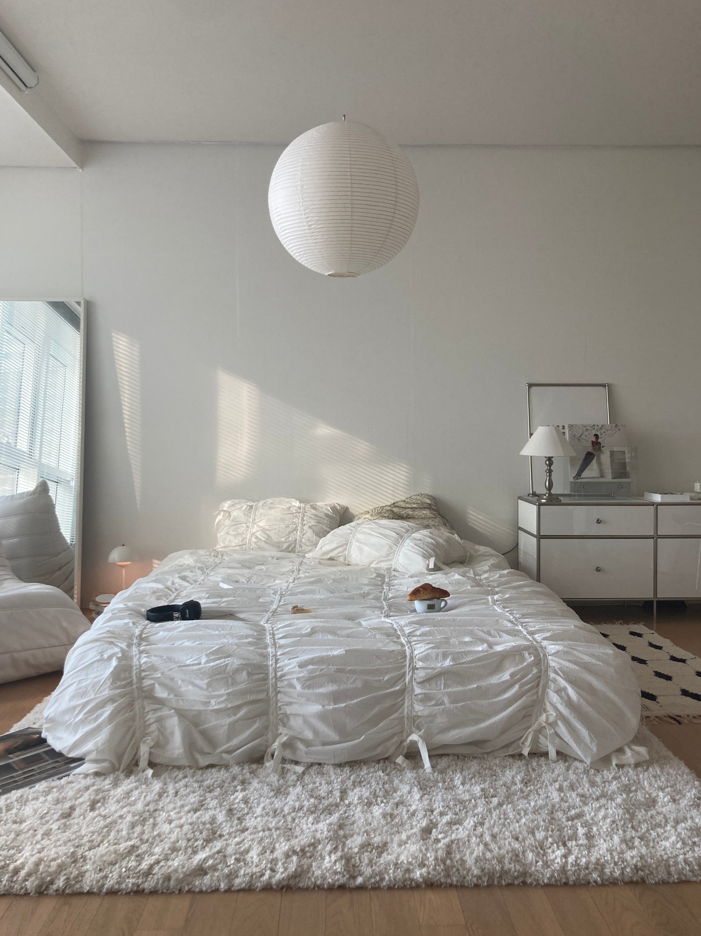 Classic white ruched bedding set in a bright all-white minimalist bedroom with a round paper pendant light and mirrored nightstand