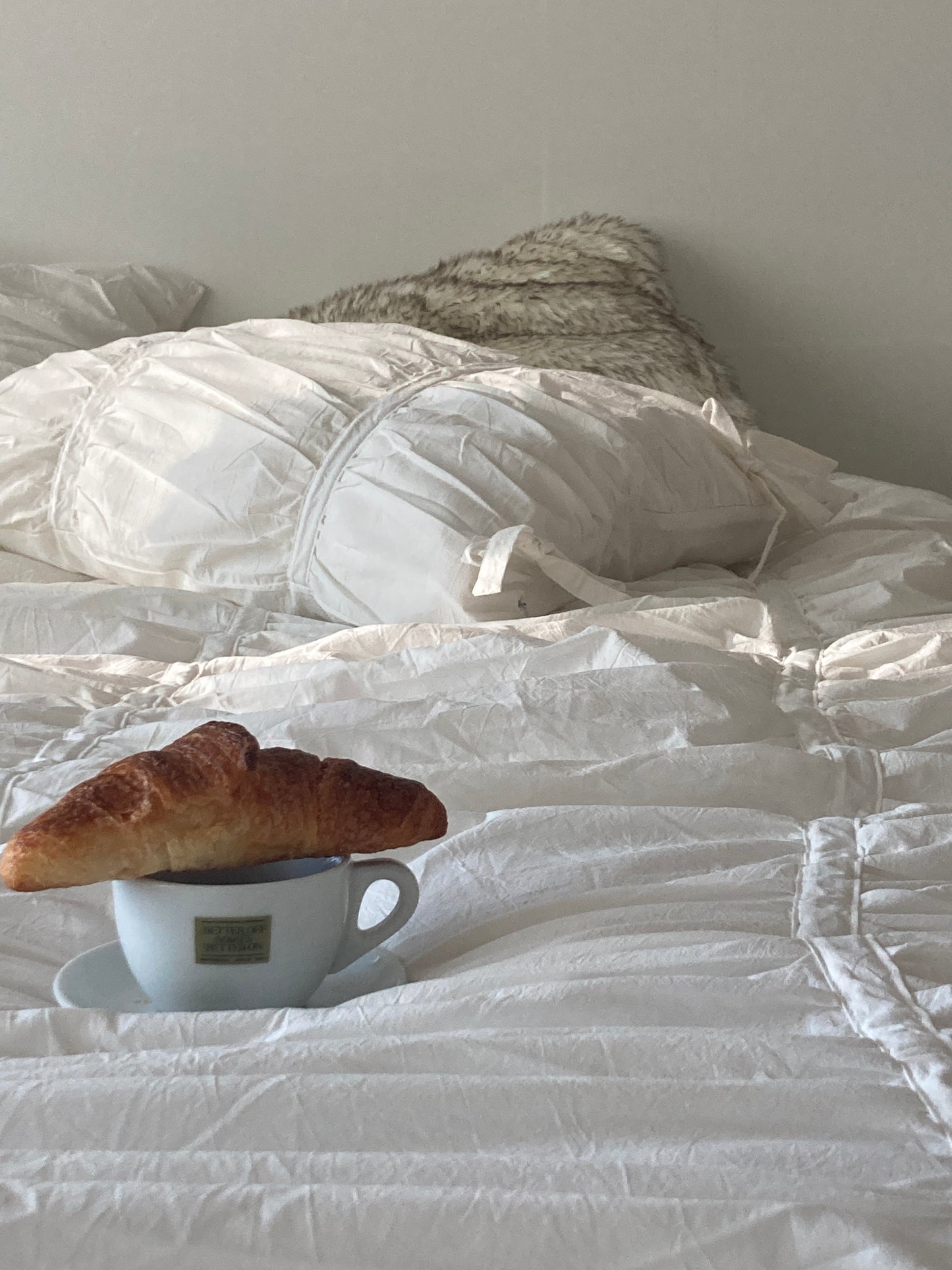 Croissant resting on a coffee cup placed on white ruched bed sheets — cozy breakfast in bed morning scene