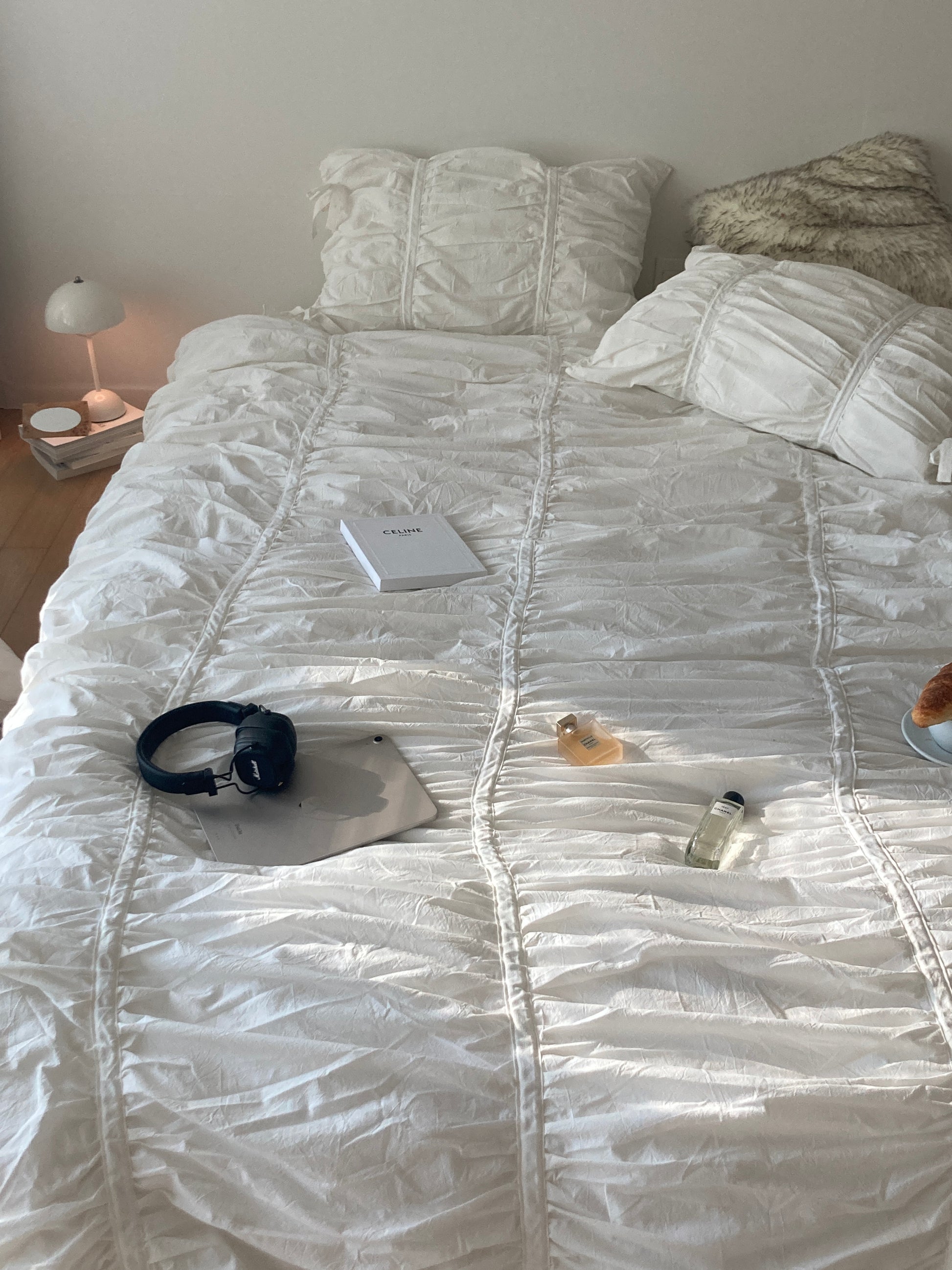 Overhead flat lay of a white ruched duvet cover set styled with a Celine book, Marshall headphones, perfume bottles, and a croissant