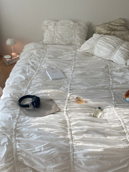 Overhead flat lay of a white ruched duvet cover set styled with a Celine book, Marshall headphones, perfume bottles, and a croissant
