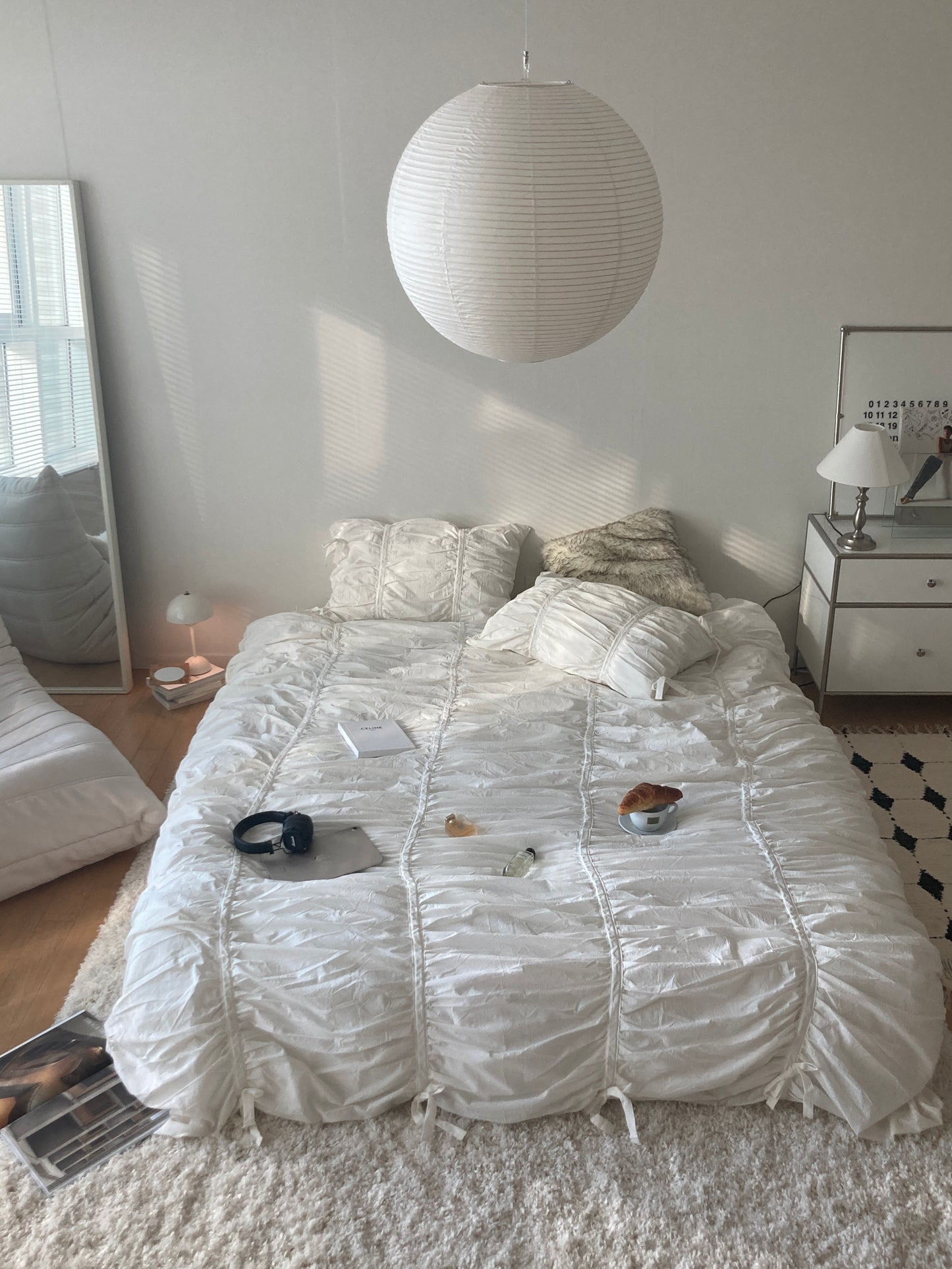 White ruched duvet cover set in a sunlit minimalist bedroom with headphones, croissant, coffee, and perfume styled on the bed